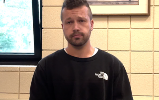 A white man with short hair and a short beard in a black crewneck sweater sitting at a table. There is a piece of paper on the table and a tile wall in the background and a partially opened window.