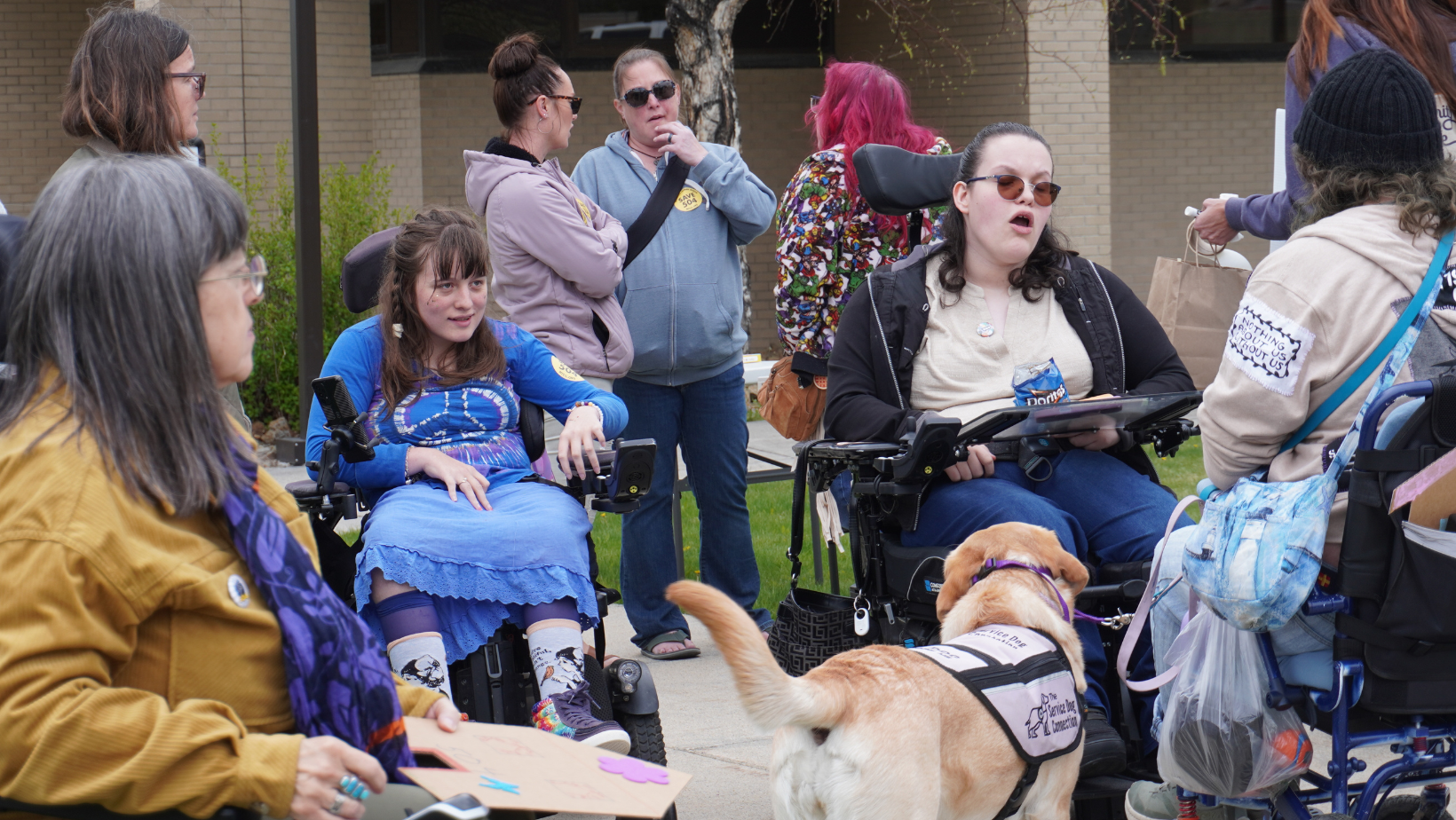 Several people with disabilities, some visible some less visible, some using chairs, some standing, and a service dog at a rally for disability rights outside the Attorney General's office in Helena MT